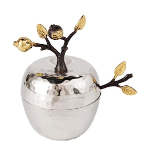 May include: A silver apple-shaped container with a hammered texture. The lid is topped with a bronze-colored branch featuring gold-colored leaves and fruit. The container is designed for decorative or functional use.