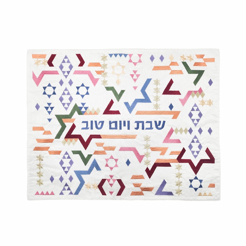 May include: A white fabric wall hanging with a geometric pattern in shades of pink, blue, green, and red. The Hebrew text "Shabbat Shalom" is embroidered in blue thread.