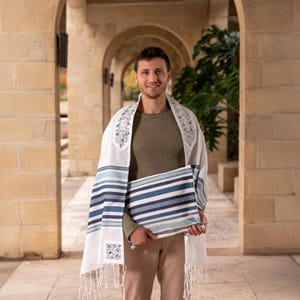May include: A man wearing a white prayer shawl with blue and white stripes and a floral pattern. He is holding a blue and white striped prayer shawl in his hands.