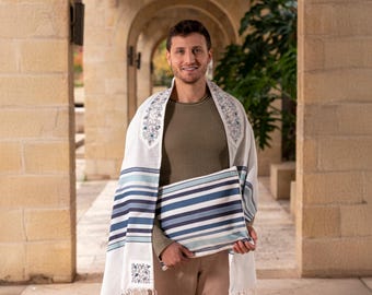 May include: A man wearing a white prayer shawl with blue and white stripes and a floral pattern. He is holding a blue and white striped prayer shawl in his hands.