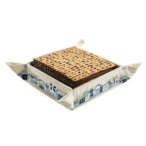 Peut inclure: Un panier carré en tissu beige rempli de matzah. Le panier présente un motif de village bleu et blanc sur les bords. La matzah est une pile de pain azyme fin avec une surface texturée.