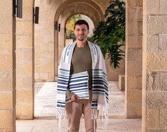 May include: A white and blue striped prayer shawl with matching carrying case. The shawl has blue stripes and text along the edges. The person is wearing a green shirt and tan pants. The background is a stone archway.