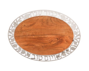 May include: An oval wooden serving tray with a silver metal rim. The rim is decorated with a Hebrew inscription and pomegranate designs.