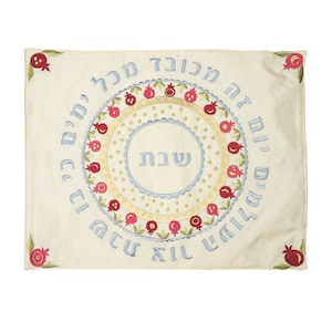 May include: A white embroidered tablecloth with a circle of pomegranates and Hebrew text. The text reads "Le'chaim" and "Shabbat Shalom".