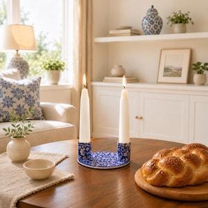 Peut inclure: Un bougeoir en céramique bleu et blanc avec deux bougies blanches allumées. Le bougeoir a un motif floral et repose sur une table en bois. Un pain est posé sur une planche en bois à proximité.