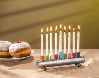 May include: A silver menorah with nine white candles lit. The candles are in colorful holders. The menorah is on a table with two donuts in the background.
