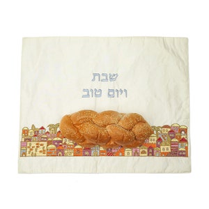 May include: A white embroidered challah cover with a braided challah on top. The cover features a border of colourful houses and the Hebrew words "Shabbat Shalom" in blue thread.