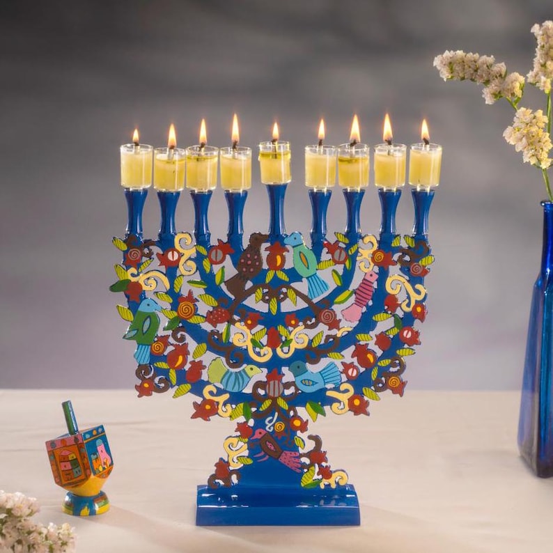 Op de afbeelding: Een blauwe menorah met brandende kaarsen, met een boomontwerp met kleurrijke vogels en bloemen. Een draaiende tol staat links. Een blauwe vaas met bloemen staat rechts.