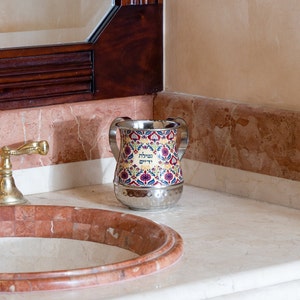 Pode incluir: Um jarro de metal decorativo com um padrão floral colorido e texto hebraico. O jarro tem uma base de prata martelada e duas pequenas alças. Está sobre uma bancada de mármore ao lado de uma pia com uma torneira dourada.