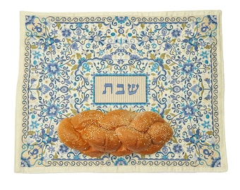 May include: A white challah cover with a blue and white floral pattern. The cover has the Hebrew word "Shabbat" embroidered in the center. A braided challah bread is placed on top of the cover.