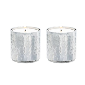 May include: Two lit candles in silver-colored, hammered metal holders. The candles have white wax and are cylindrical, with a small wick visible. The holders have a textured surface and a simple, modern design.