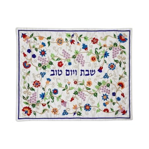 May include: White embroidered fabric with a floral design and the Hebrew text "Shabbat Shalom" in blue.