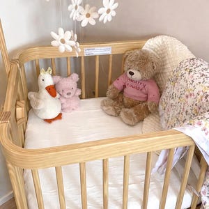 May include: A light wood baby crib with a white mattress, holding stuffed animals. A white daisy mobile hangs above. The crib contains a white goose with a crown, a pink teddy bear, and a brown teddy bear wearing a pink shirt.