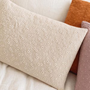 May include: A rectangular beige pillow with a floral pattern made of small white embroidered flowers. The pillow is on a white bed with other pillows in shades of orange and pink.