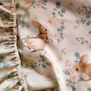 May include: A cream-colored fabric with a floral and bunny print. The design features watercolor-style illustrations of bunnies, flowers, and foliage in soft pastel colors. One bunny sits in a teacup. The fabric has a ruffled edge.