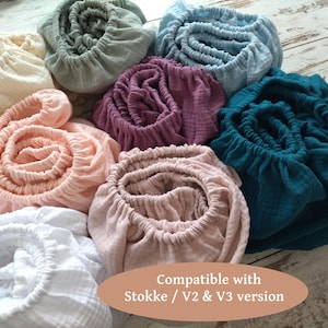 May include: A selection of fitted cot sheets in a range of colours: cream, grey, blue, purple, pink, and teal. The sheets are made from a soft, textured material and have elasticated edges. Text on the image reads: "Compatible with Stokke / V2 & V3 version."