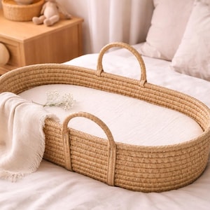 May include: Woven wicker baby bassinet with handles, featuring a white interior and a cream-colored blanket. The oval-shaped bassinet is placed on a bed with a white sheet. A small sprig of white flowers rests inside.