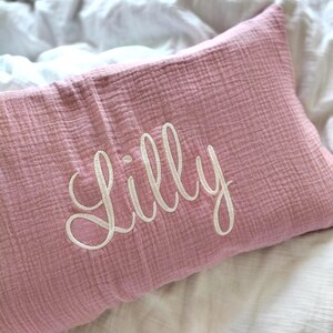 May include: A rectangular dusty pink muslin cushion featuring the name Lilly embroidered in white cursive script across the centre. The fabric has a distinct crinkled texture characteristic of multi-layered cotton gauze, providing a soft appearance.