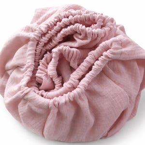 Pode incluir: Um lençol de elástico macio em rosa seco, feito de um tecido texturizado e enrugado. O lençol apresenta uma borda elástica franzida projetada para prender ao redor de um colchão, criando uma aparência arredondada com dobras e textura visíveis.