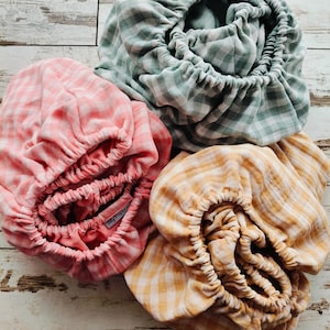 Op de afbeelding: Vier hoeslakens voor wiegjes in pastelkleuren: roze, geel en groen, elk met een ruitpatroon. De lakens zijn aan de randen gerimpeld met elastiek. De lakens zijn gemaakt van zachte stof.