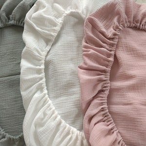 May include: Three fitted sheets made of crinkled muslin fabric, each featuring an elasticized border. The sheets are presented in three distinct solid colors: a muted sage green, a crisp white, and a soft dusty rose pink.
