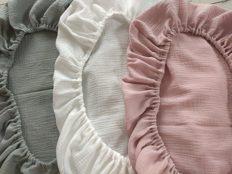 May include: Three fitted sheets made of crinkled muslin fabric, each featuring an elasticized border. The sheets are presented in three distinct solid colors: a muted sage green, a crisp white, and a soft dusty rose pink.