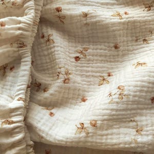 May include: Cream-coloured fitted sheet with a delicate floral pattern. The fabric has a crinkled texture, and the flowers are in shades of brown and red. The elasticated edge is visible on the left side of the image.