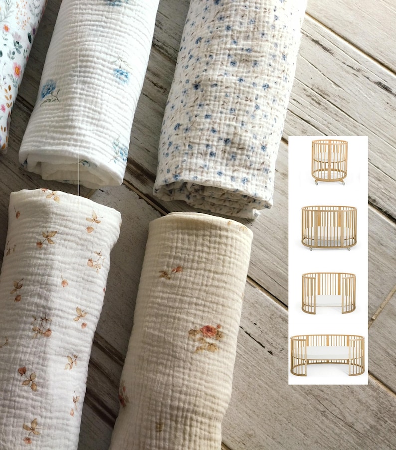 May include: Rolled muslin baby blankets with floral patterns in white, cream, and blue. The blankets are displayed on a light wood surface. A crib is shown in the corner, with a round design and wooden frame.