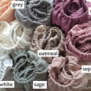 May include: A collection of nine gathered fabric items in various colors labeled as peach, grey, wild berry, cream, oatmeal, dusty pink, white, sage, and sepia rose. Each piece features a crinkled texture and an elasticized opening for a fitted look.