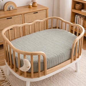 May include: A light wood baby crib with a sage green fitted sheet. The crib has a unique curved design with a white base and legs. A hanging moon-shaped mobile adds a decorative touch. The crib is on a cream-colored rug.
