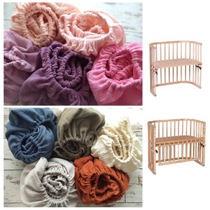 May include: A collection of gathered, elasticized fabric sheets in various colors including peach, purple, pink, blue, olive, cream, and rust, displayed alongside two wooden cribs with slatted sides and adjustable height platforms.