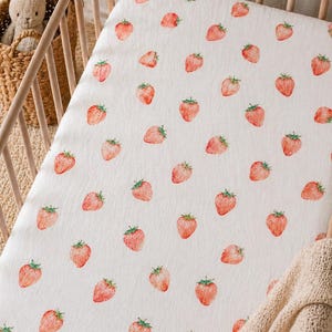 May include: A white cot sheet featuring a repeating pattern of small, watercolour-style red strawberries with green leaves. The fabric is stretched over a mattress within a wooden cot frame, with a woven basket and a plush toy visible nearby.