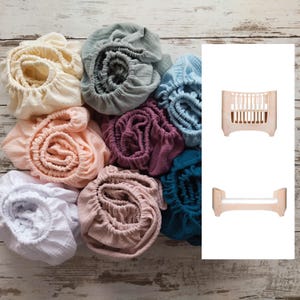 May include: A selection of rolled-up cot bed sheets in pastel shades such as cream, peach, and blue. A light-colored wooden cot bed is shown in two views, top and side.