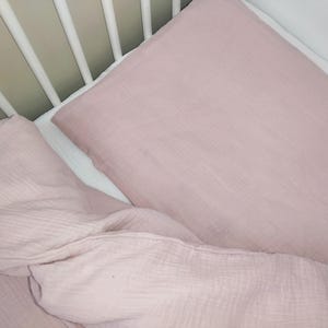 May include: Two pink muslin crib sheets, one folded and one unfolded, in a white crib.