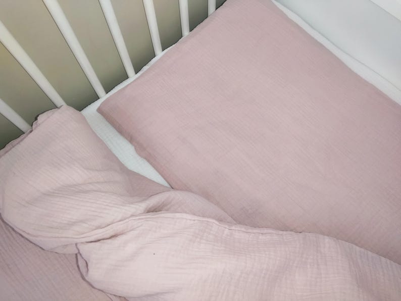 May include: Two pink muslin crib sheets, one folded and one unfolded, in a white crib.