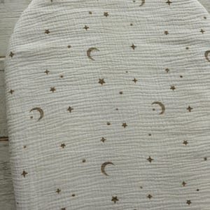 May include: Off-white, oval-shaped baby changing pad cover with a celestial pattern. The design features brown crescent moons, stars, and small dots scattered across the surface. The fabric has a textured, crinkled appearance.