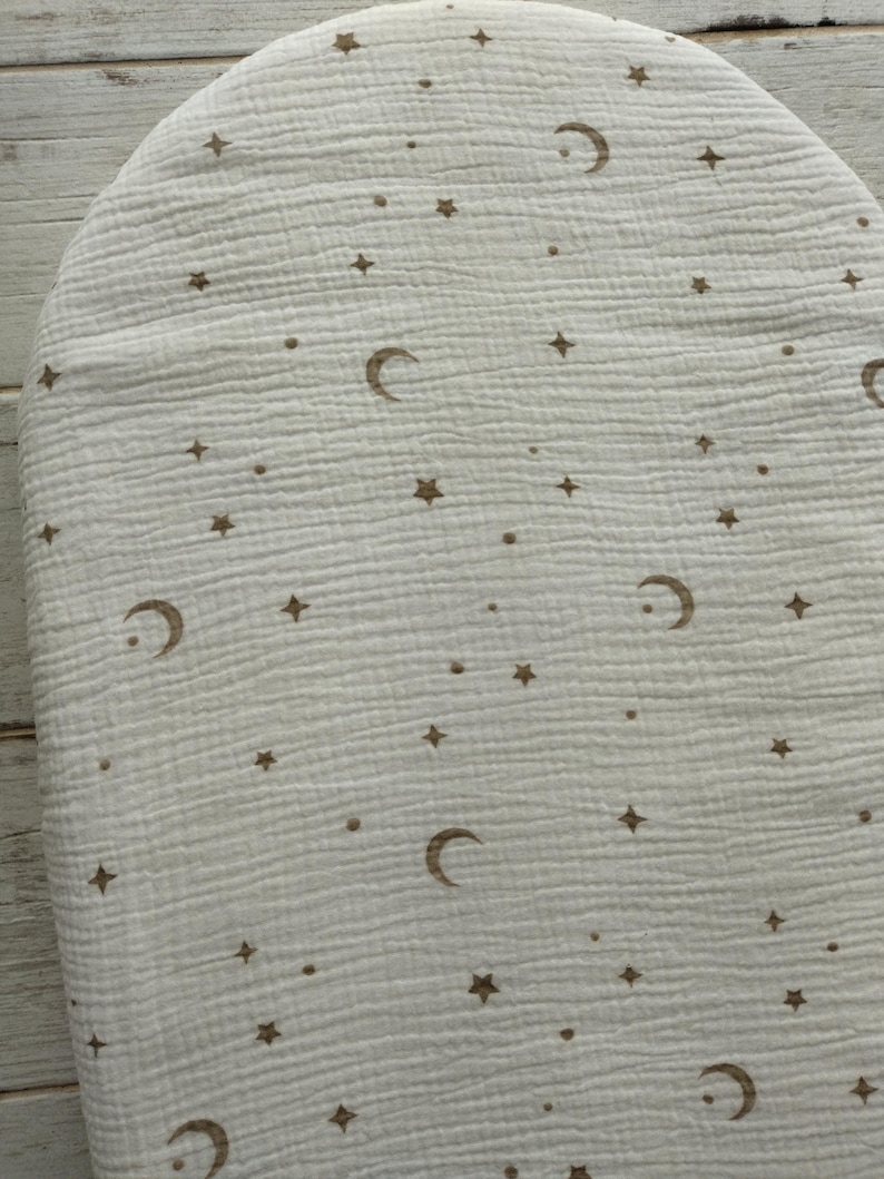 May include: Off-white, oval-shaped baby changing pad cover with a celestial pattern. The design features brown crescent moons, stars, and small dots scattered across the surface. The fabric has a textured, crinkled appearance.