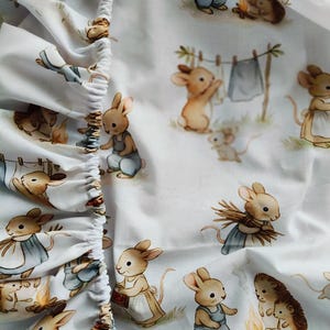 May include: A white fitted sheet featuring a repeating pattern of illustrated brown rabbits and hedgehogs. The rabbits are depicted wearing blue overalls or aprons, performing tasks like hanging laundry on a line or gathering wood near a small campfire.