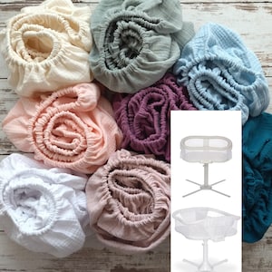 Op de afbeelding: Een verzameling hoeslakens voor wiegjes in verschillende pastelkleuren, waaronder crème, roze, paars en blauwgroen. De lakens zijn gemaakt van een zachte, getextureerde stof en zijn in een cirkelvormig patroon gerangschikt. Een witte wieg is ook afgebeeld.