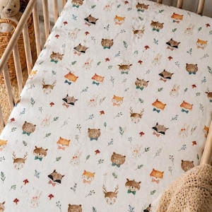 May include: A white cot sheet featuring a repeating pattern of watercolour forest animals including bears, foxes, deer, and rabbits, each wearing a small bow tie, interspersed with tiny green sprigs and small red mushrooms across the fabric.