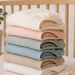 May include: A stack of six folded muslin crib sheets in solid colors including cream, soft pink, sage green, dusty blue, beige, and white. Each sheet features a textured, crinkled fabric surface and elasticized edges for a secure fit on a mattress.