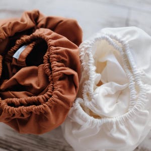 Op de afbeelding: Close-up van twee hoeslakens voor wiegjes. Eén is een warme bruine kleur en de andere is zacht wit. Beide lakens hebben elastische randen, waardoor ze een gerimpeld uiterlijk krijgen. De lakens zijn gemaakt van een zacht, mogelijk katoenen materiaal.