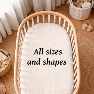 Può includere: Una culla ovale in legno con sbarre verticali bianche e un coprimaterasso testurizzato color crema. Al centro del materasso è presente il testo nero All sizes and shapes stampato con un carattere serif chiaro, centrato sulla superficie.