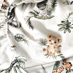 May include: A white fitted sheet featuring a repeating pattern of watercolour-style jungle animals including a lion cub, a giraffe, and a hippopotamus, interspersed with various green tropical palm leaves and foliage throughout the fabric.