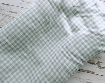 Op de afbeelding: Een zachte, lichtgewicht stoffen deken met een klassiek saliegroen en wit gingham ruitpatroon. Het textiel heeft een licht gekreukeld, getextureerd uiterlijk met een consistent rasterontwerp over het gehele oppervlak van het gevouwen materiaal.