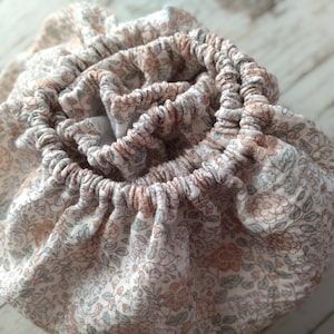 May include: A white fabric pouch with a floral pattern in shades of brown and green. The pouch is gathered at the top and has a ruffled edge.