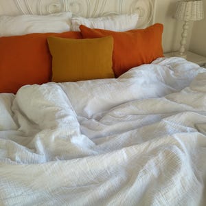 May include: A white bed with a white duvet cover and three pillows. The pillows are covered in orange and yellow fabric.