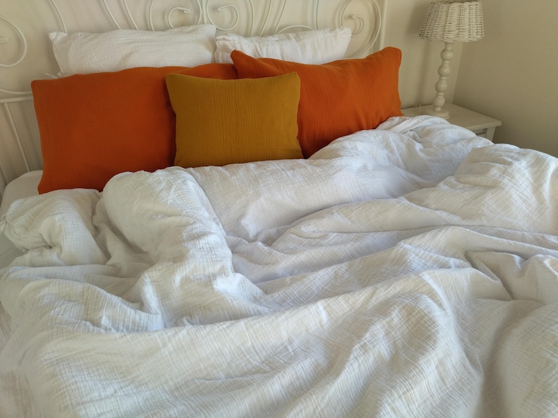 May include: A white bed with a white duvet cover and three pillows. The pillows are covered in orange and yellow fabric.