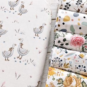 Può includere: Un lenzuolo in tessuto bianco con un motivo ripetuto di oche grigie con nastri marroni, accanto a una pila di cinque tessuti arrotolati con stampe varie tra cui forme astratte, rose rosa, rametti botanici e motivi floreali gialli.