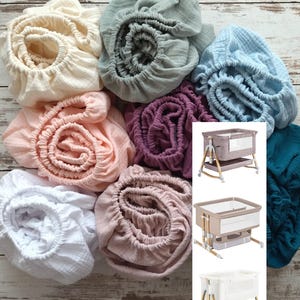 May include: A collection of fitted crib sheets in various colors, including cream, gray, blue, pink, purple, and white. The sheets are made of a soft, textured fabric. Three baby cribs are also shown, each with a different color scheme.